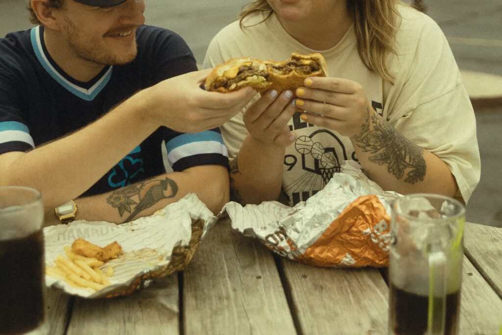 a couple cheers their burgers as they sit at an outdoor wooded table