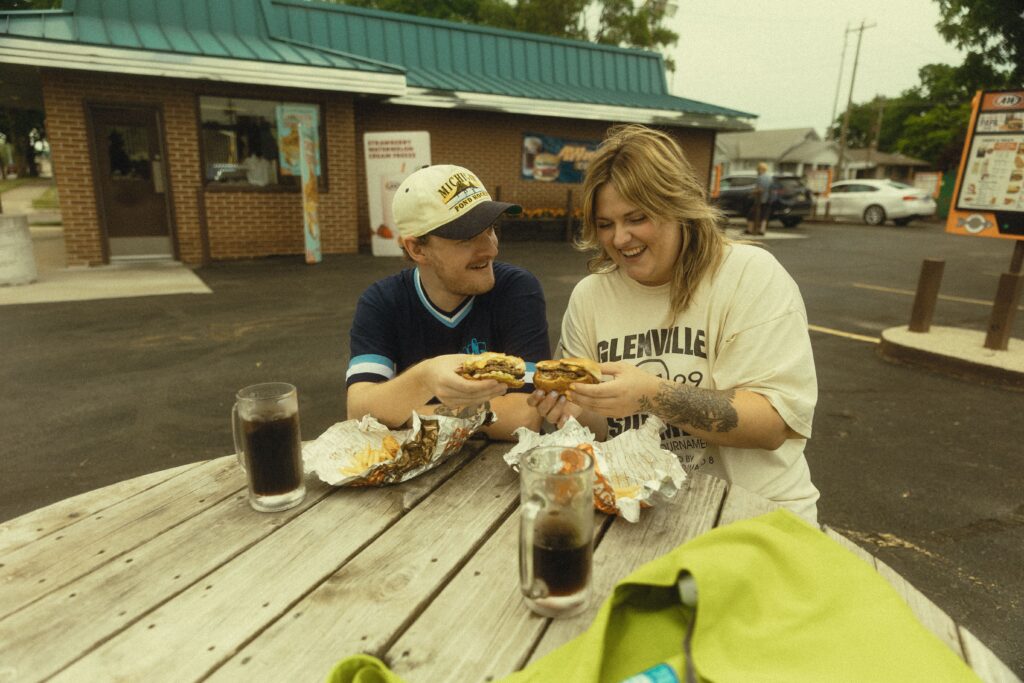 a couple laugh together as they eat burgers together outside