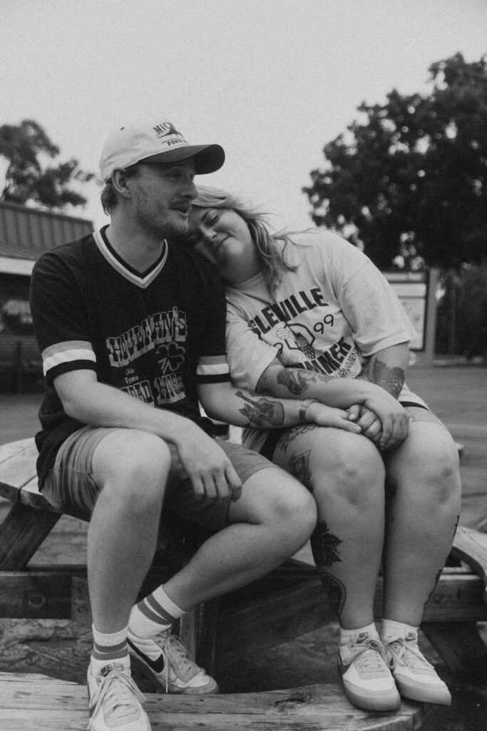 couples photography where a couple smile together while sitting on an outdoor wooden table, she rests her head on his shoulder