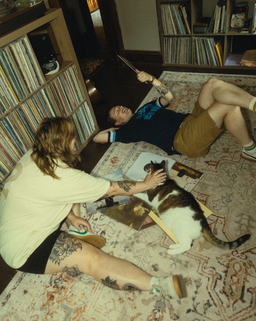 a couple sit on the floor of their living room with their cat and vinyl records