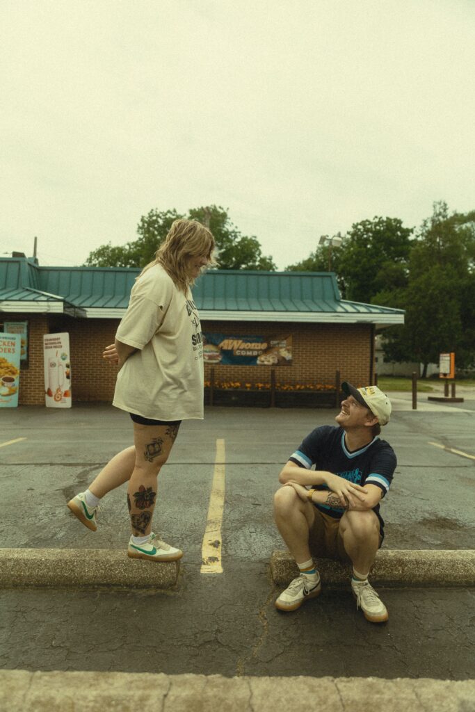 a woman stands on a parking barrier and looks down at her partner who is sitting on one beside her and smiling up at her during their couples photos