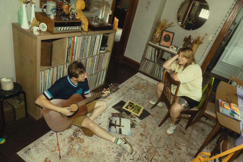 a couple sit in their home and smile at each other while he plays guitar