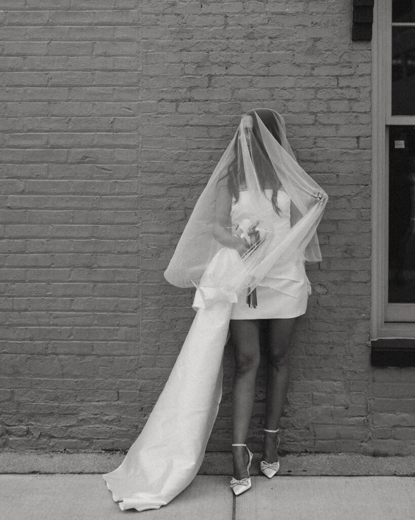 a woman wearing a short white wedding dress with a train and a veil stands in front of a brick wall during her elopement