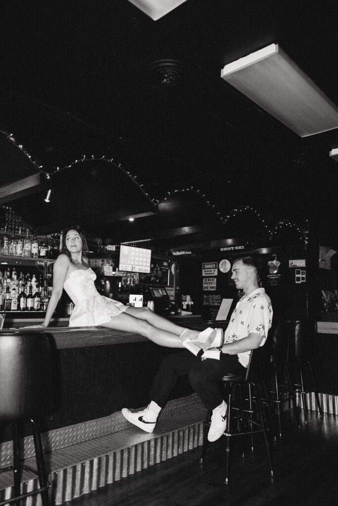 a woman in a small white dress sits on a bar while her husband sits on a chair at it