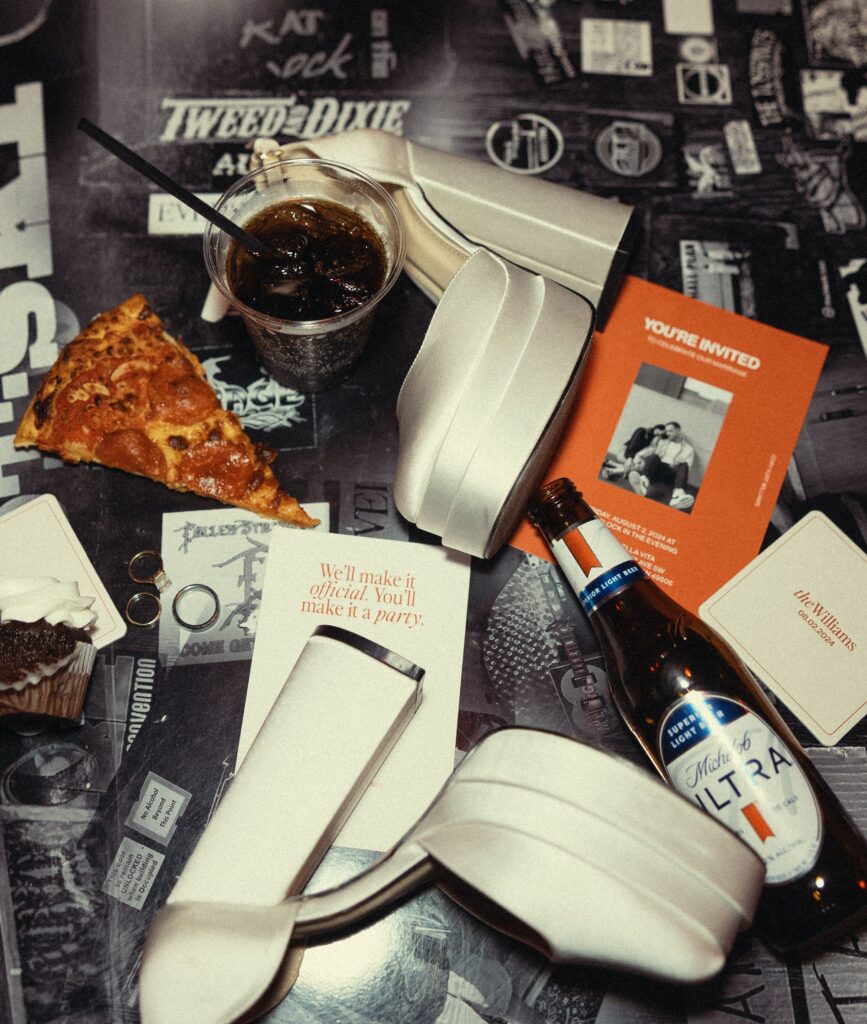 an elopement flatlay including a piece of pizza, a beer, a mixed drink, white heels, rings, and invitations