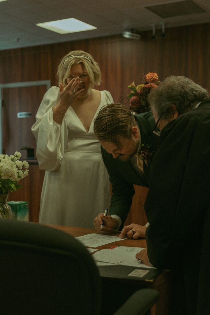 how to elope in michigan a couple wearing wedding attire smile as they sign documents in a courthouse