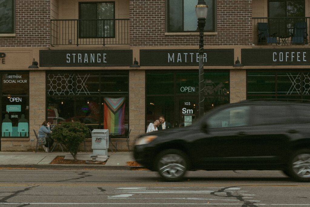 a couple sit across the street in front of a coffee shop during their elopement as a car passes by