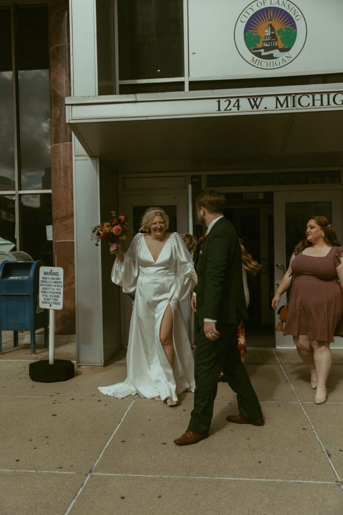 a couple eloping in michigan wearing wedding addire smile outside of the city of lansing courthouse