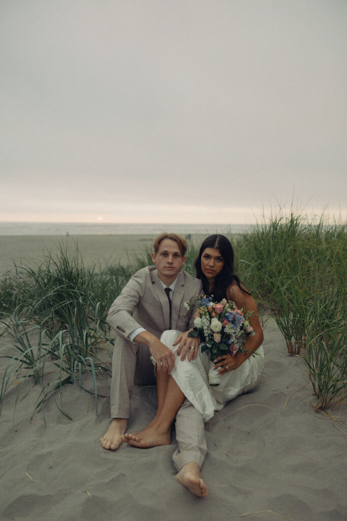 a couple in wedding attire sit on the beach during elopement photography for their wedding