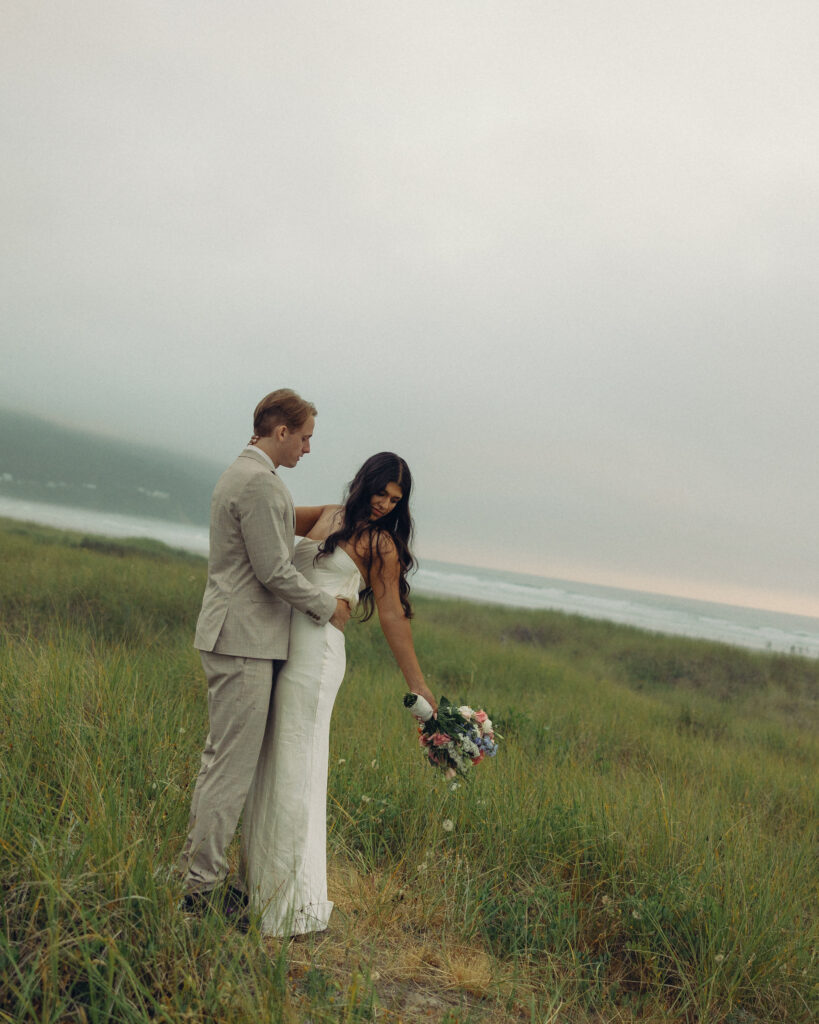 a couple stands in a field in front of a beach during their oregon elopement