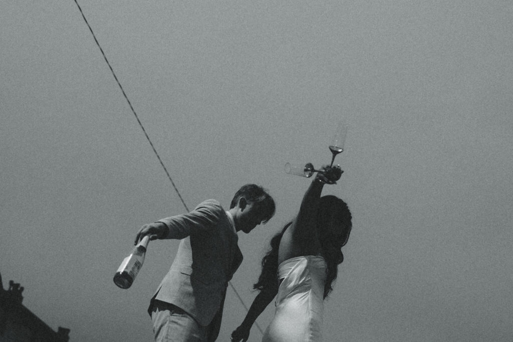 a couple walk in a line with their arms out to keep their balance, he holds a bottle while she holds two glasses during their elopement photography