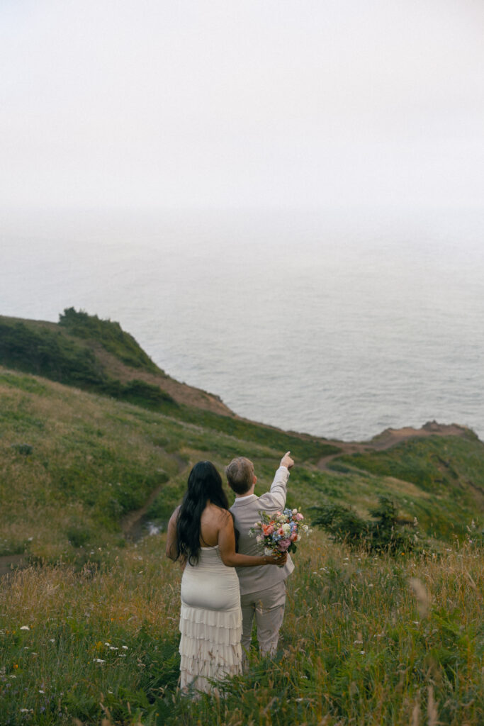 a couple stand together on a mountain in front of the ocean, he points to something in the vast distance