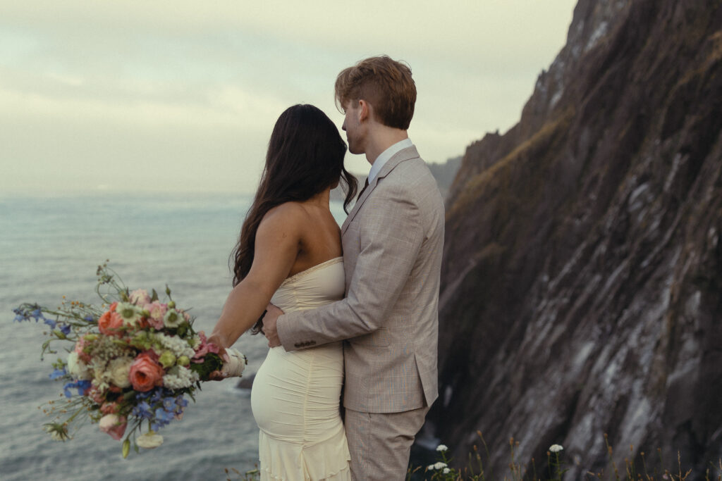 elopement photography an eloping couple stand on a mountain in front of the ocean