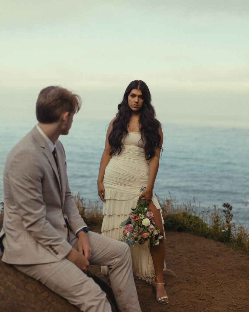 a woman stands looking directly at the camera in her wedding dress for elopement photography as her husband looks over at her