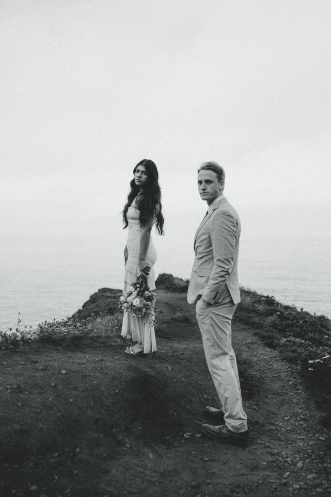 a couple look over their shoulders as they stand on a mountain in front of the ocean