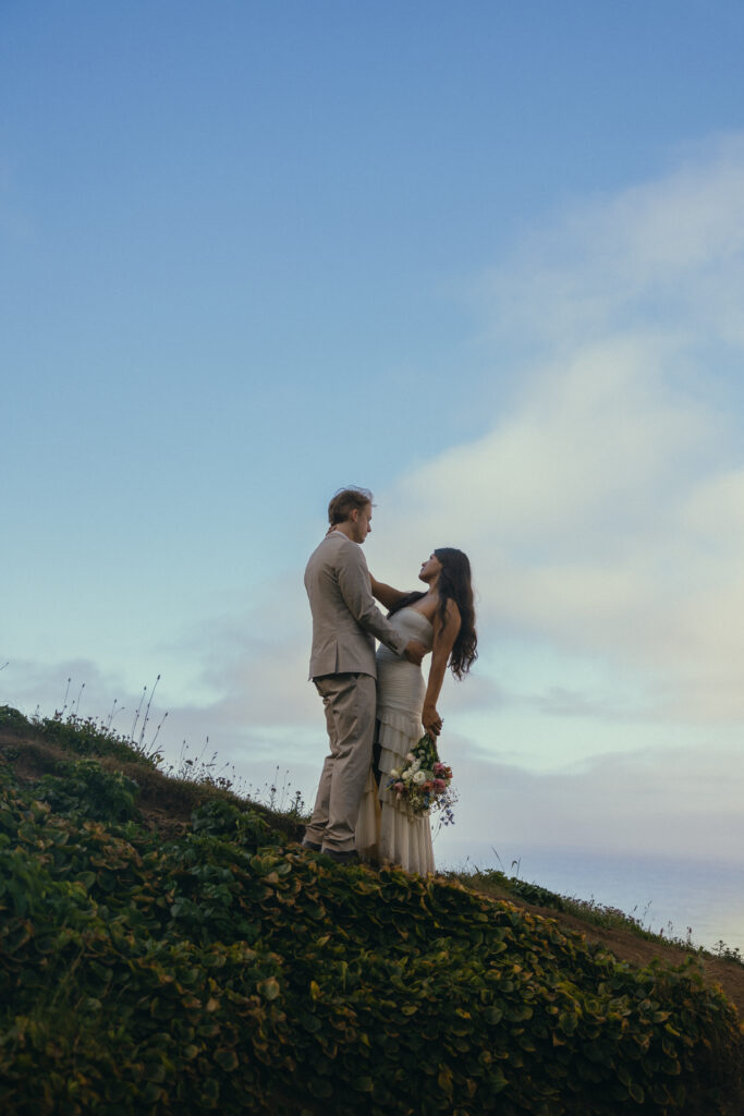 a couple gaze at each other on a hill with the blue sky behind them