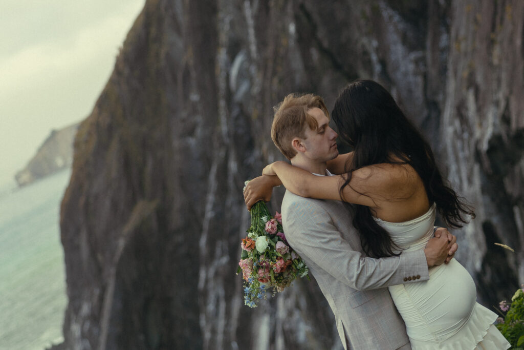 elopement photography where an eloping couple stand in front of the ocean and a mountain with her arms around his neck