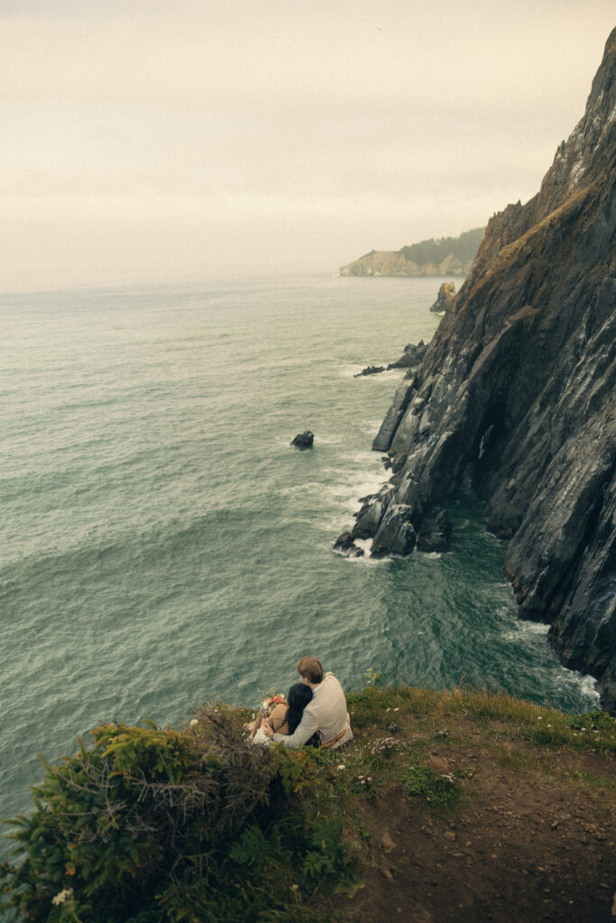 a couple sit together on the side of a cliff overlooking the ocean and the oregon mountains during their elopement