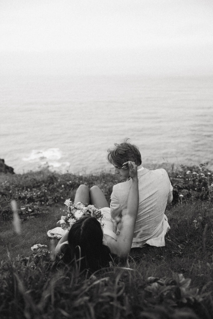 a couple sits on the ground in front of the ocean during their elopement