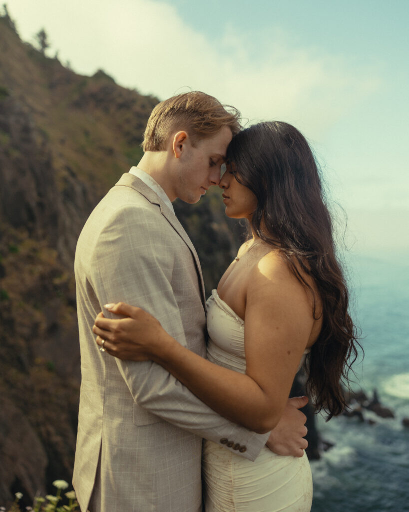 a couple lean against each other in front of the side of a mountain and the ocean