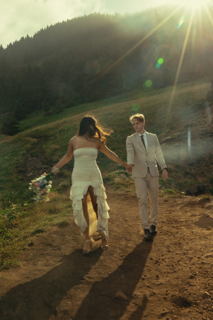 during elopement photography a couple walk hand in hand along a dirt path with a forest behind them