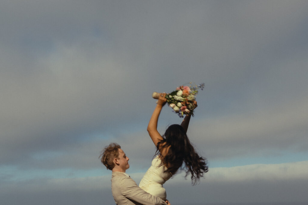 a man lifts his wife up as she waves her bouquet in the air for their elopement photography