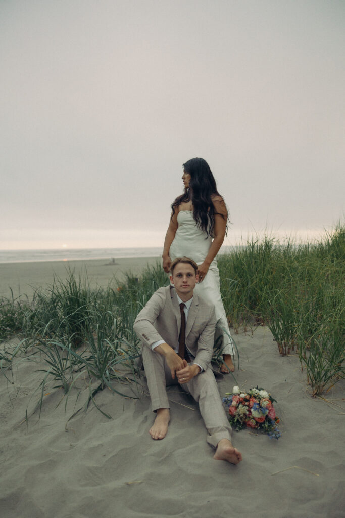 a man sits on the beach barefoot with his wife behind him during their oregon elopement