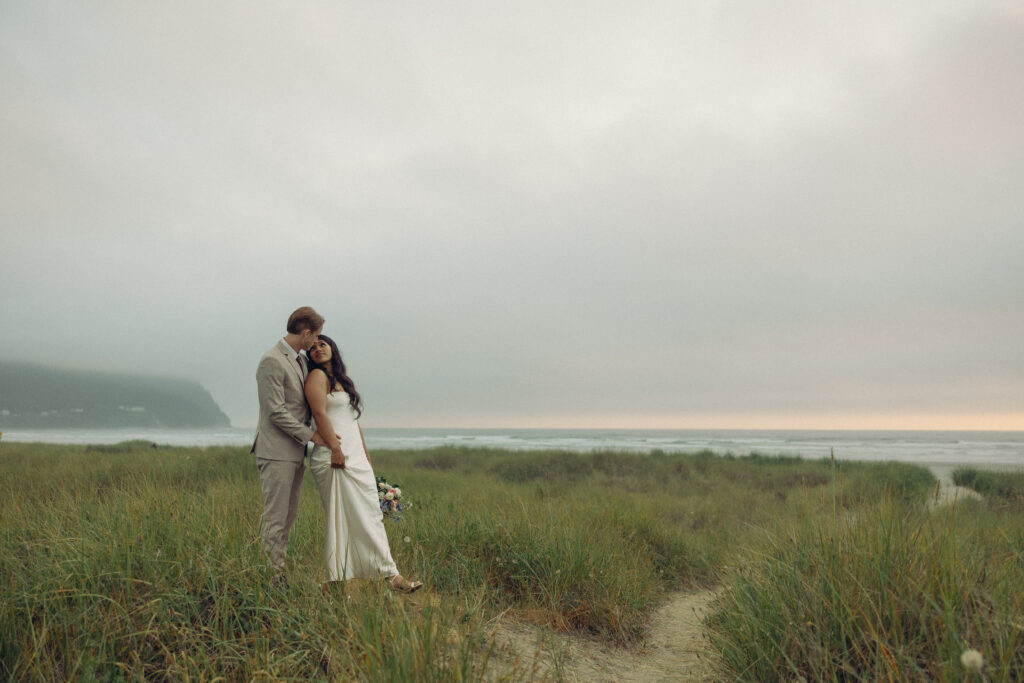 a couple stands on a west cost beach leaning against each other during elopement photography