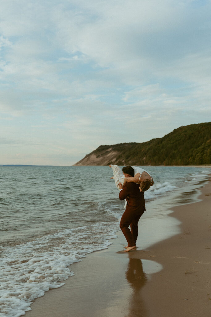 A groom throws his bride over his shoulder on a shoreline at a Michigan beach, one of the most beautiful  places to elope in michigan