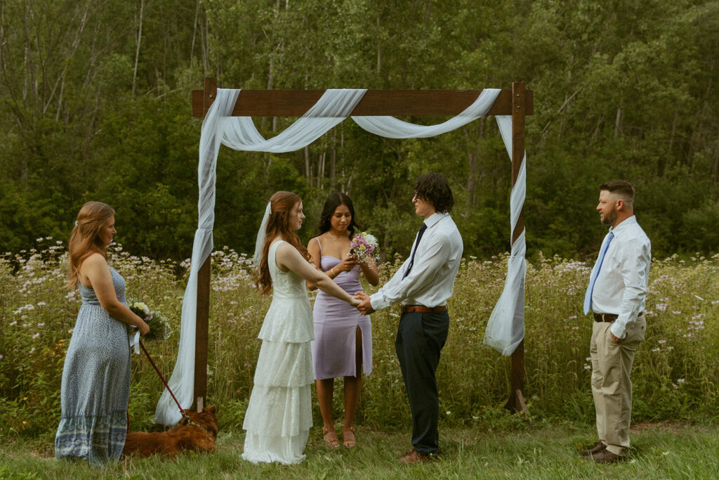 A MI couple holds hands as they exchange vows under a wooden arch at their outdoor ceremony eloping in Michigan