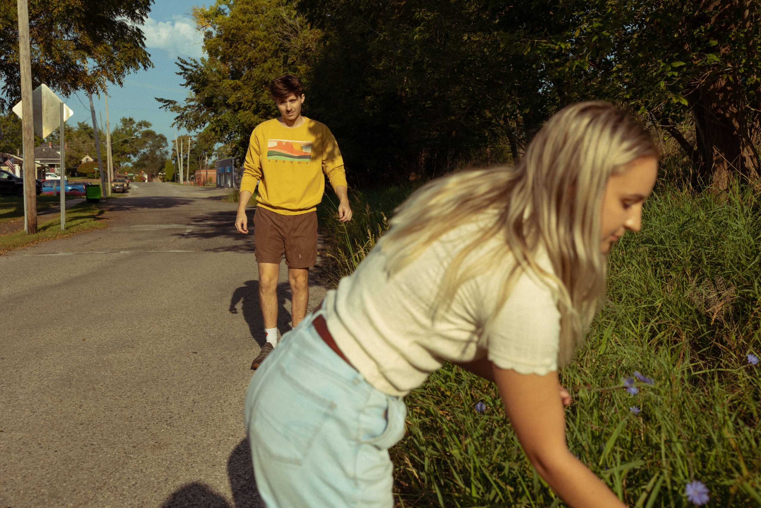 a woman leans down to pick a flower during her engagement photos as her fiancee walks towards her in the background