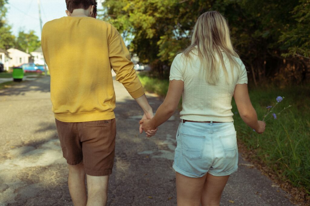 engagement photographer photo of a couple holding hands on a walk while she holds a flower she picked