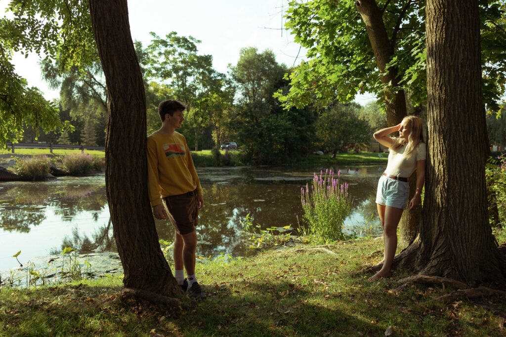 engagement photographer photo of a couple leaning against opposite trees in front of a pond