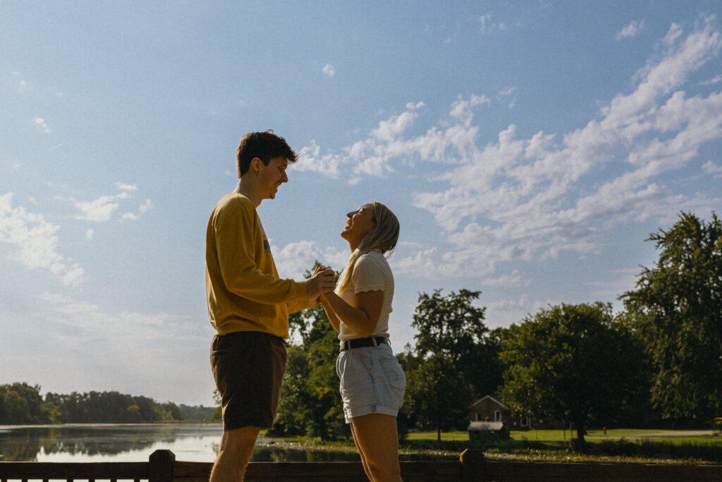 a couple stand in front of a lake facing each other laughing and holding hands for their engagement photographer