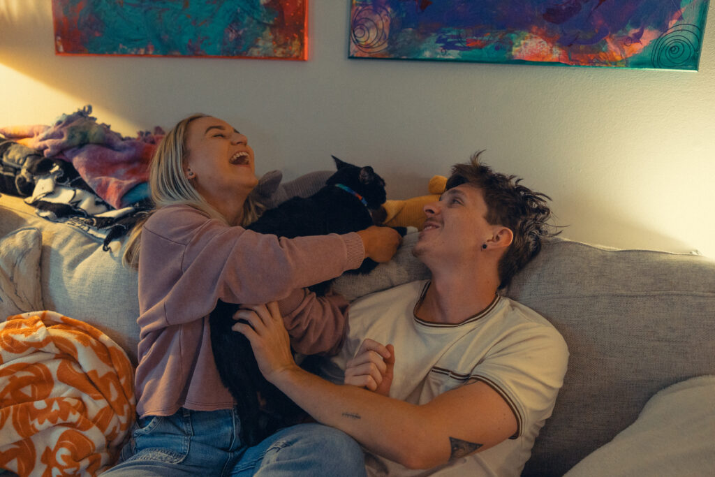 an engaged couple laugh together on a couch with their cat