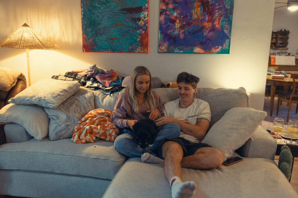 an engaged couple smile together on the couch with their cat during engagement photos grand rapids