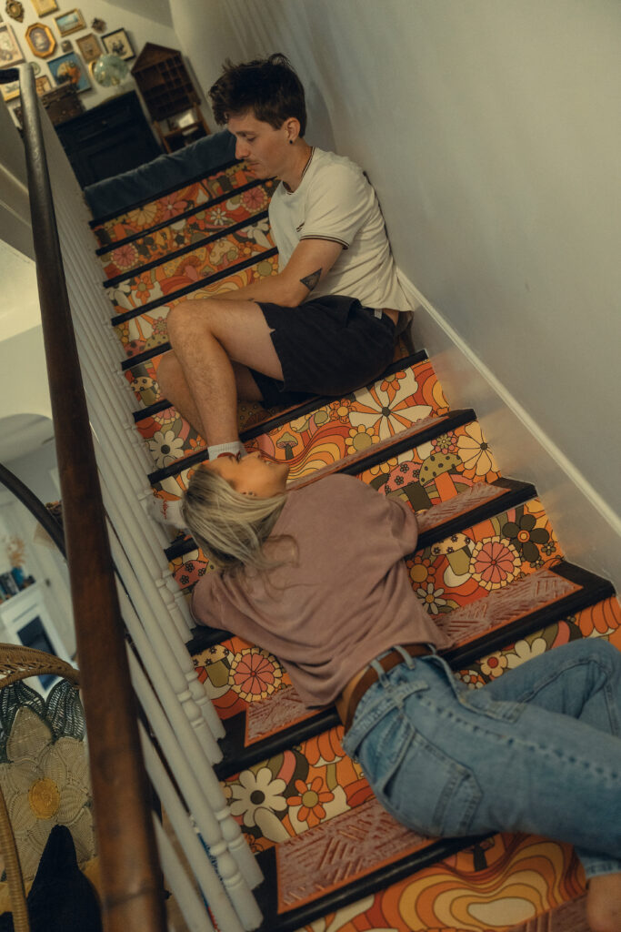 a woman sits on the stairs of her home and looks up at her fiance above her during their engagement photos grand rapids