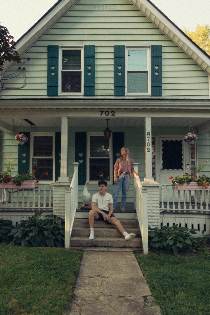 a man sits on the front step of their house with his leg out and his fiance standing behind him