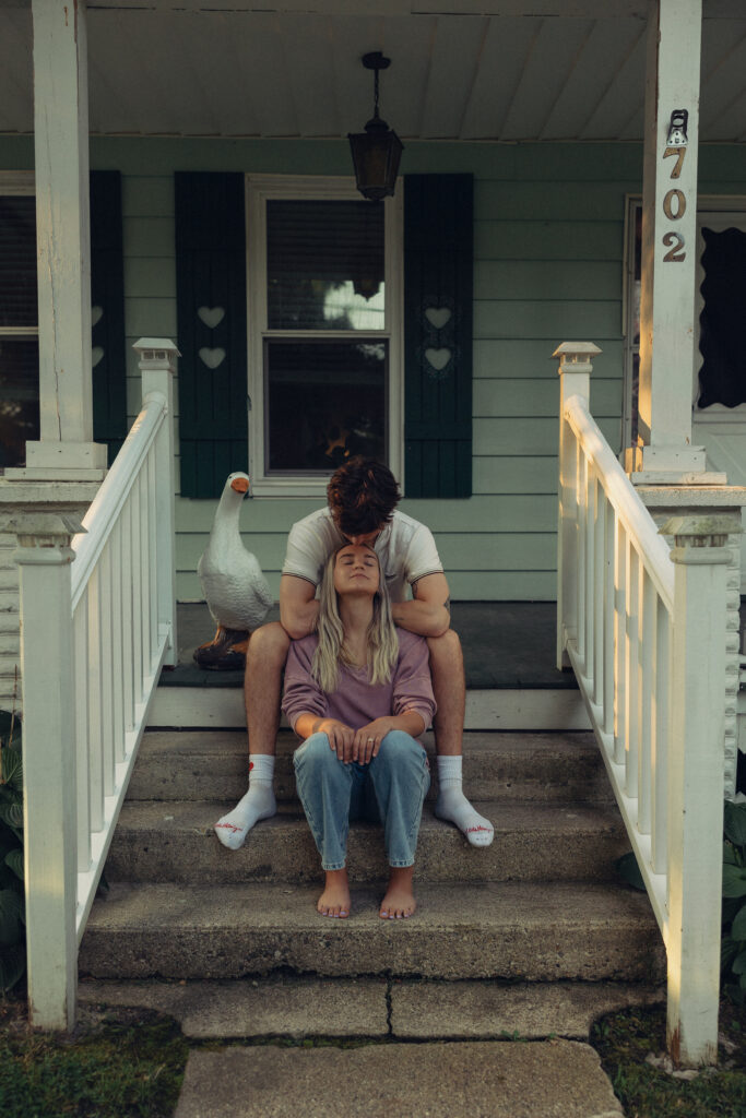 a man presses a kiss to his fiance's forehead while they sit on their front step during a session with an engagement photographer