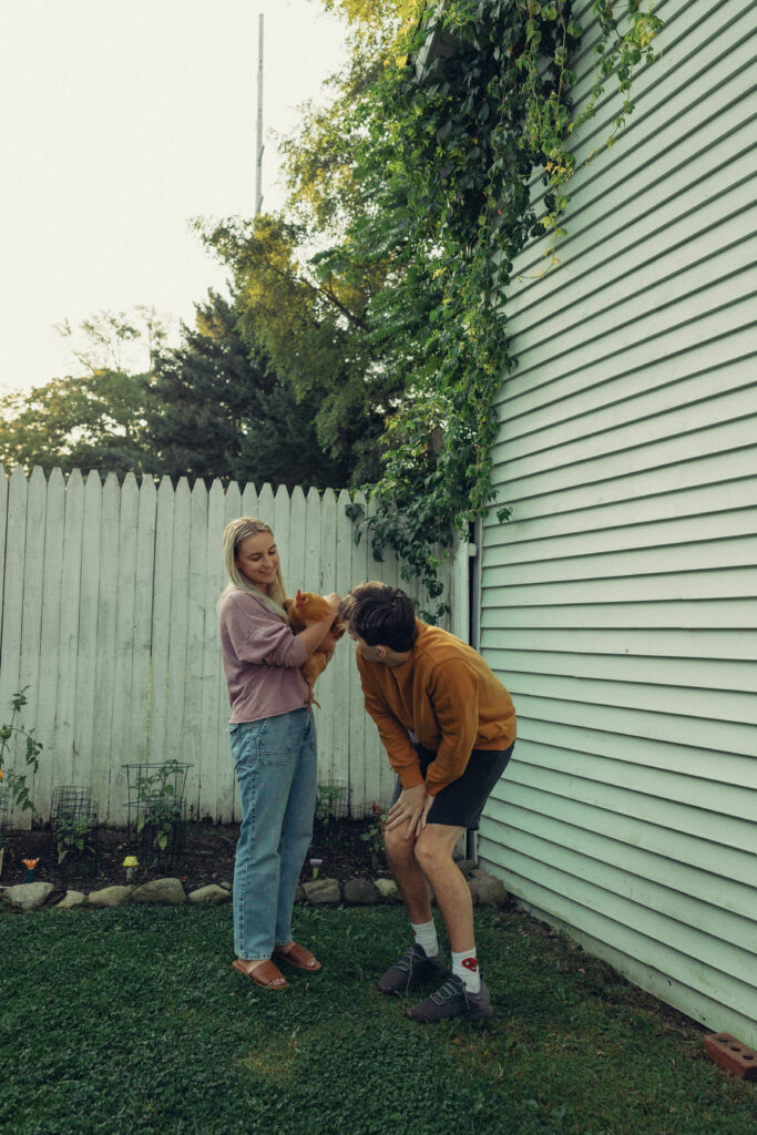 a woman holds a chicken while her fiance bends down to look at at during their engagement photos grand rapids