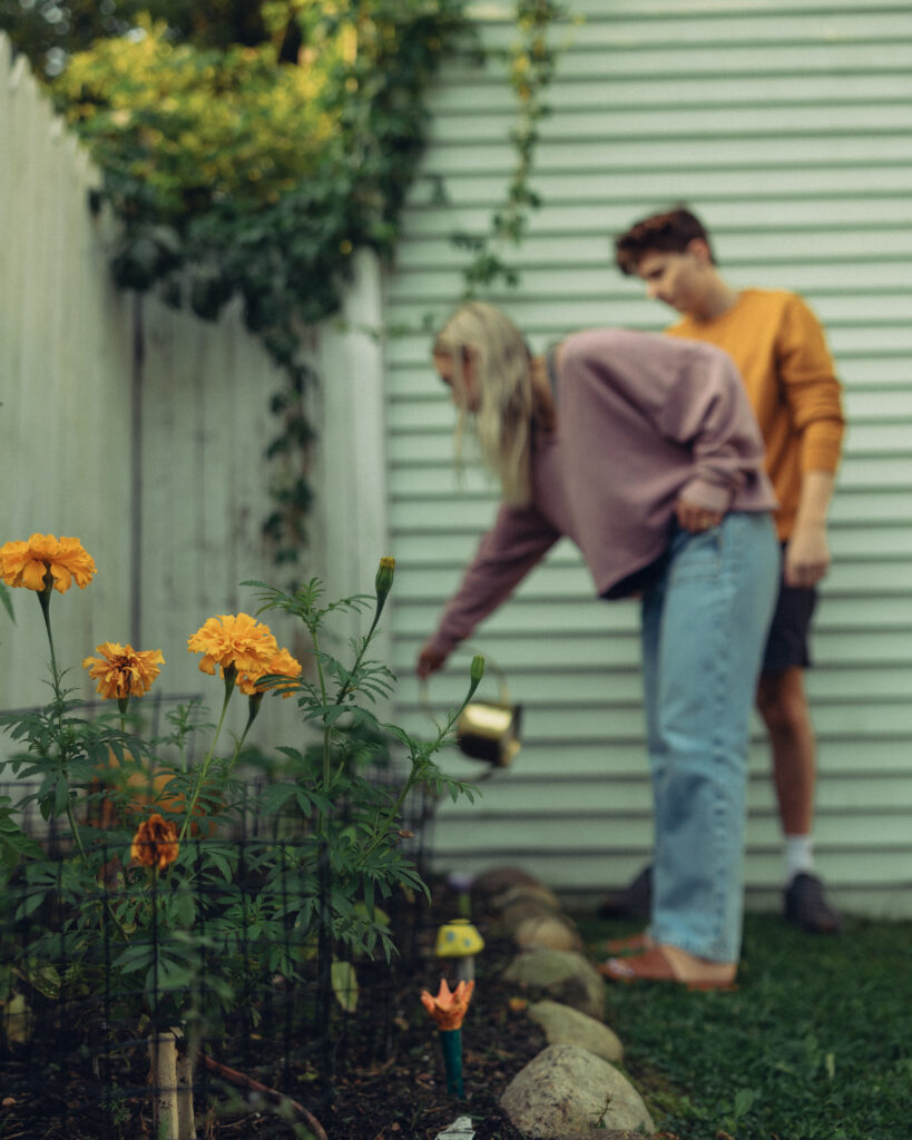 growing flowers in the foreground while an engaged couple waters plants in the background during their engagement photos grand rapids