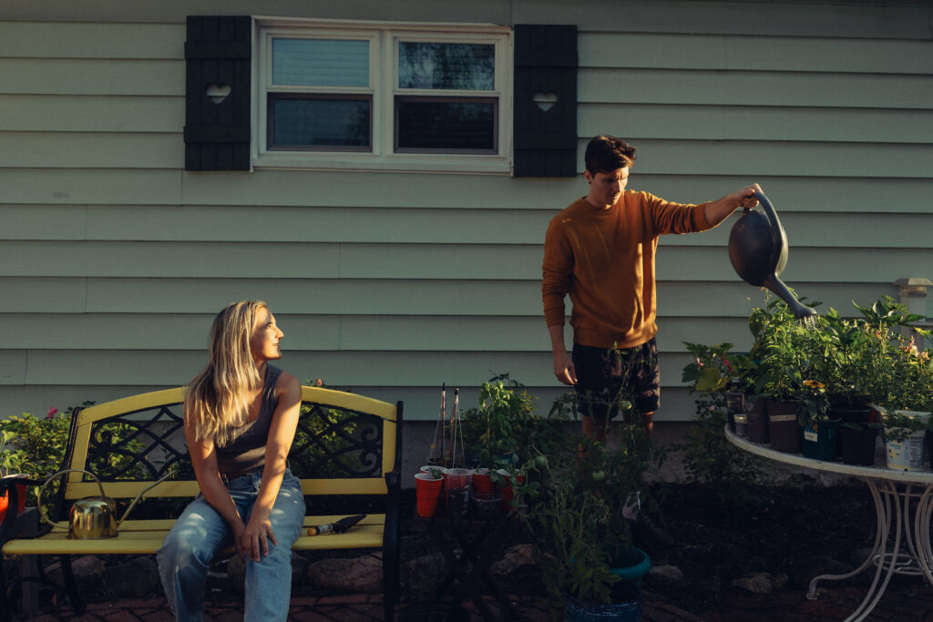 a woman looks over at her fiance who is watering plants during a session with their engagement photographer