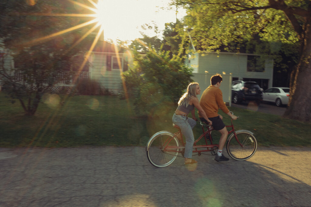 an engaged couple ride a tandem bike together with the sun flaring through trees behind them during their engagement photos grand rapids