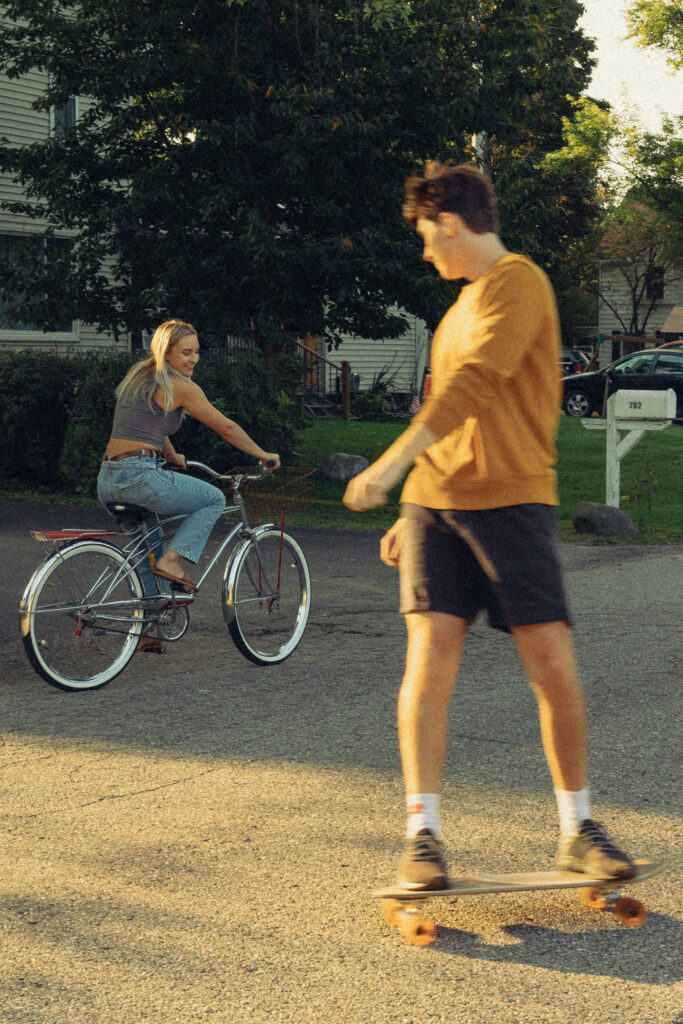 a man rides a skateboard while his fiance rides a bike behind him during a session with their engagement photographer