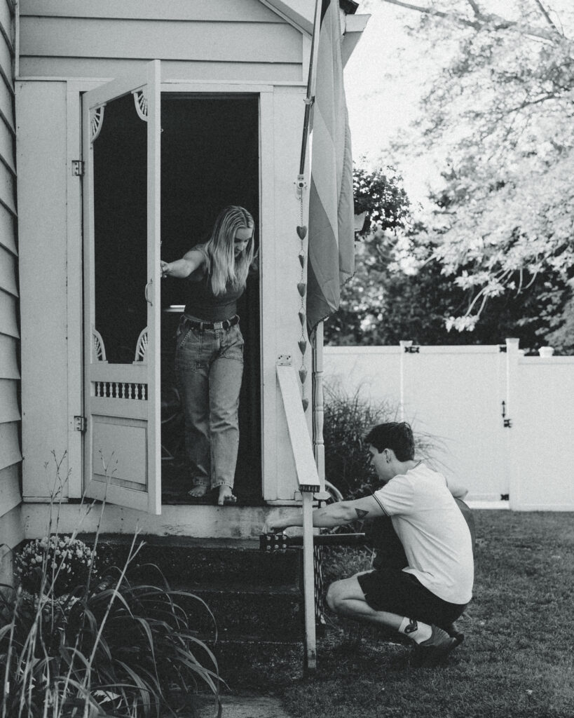 a man plays guitar while crouching on the ground as his fiance walks out the door next to hime during their engagement photos grand rapids