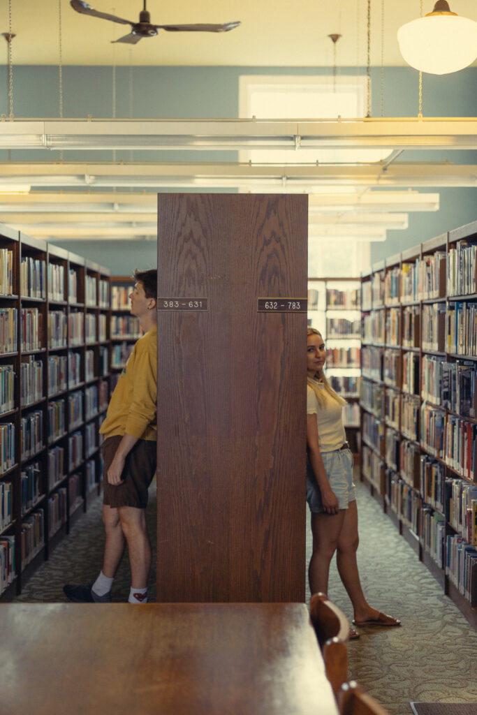 a couple stands on two sides of the same shelf at a library during their session with an engagement photographer