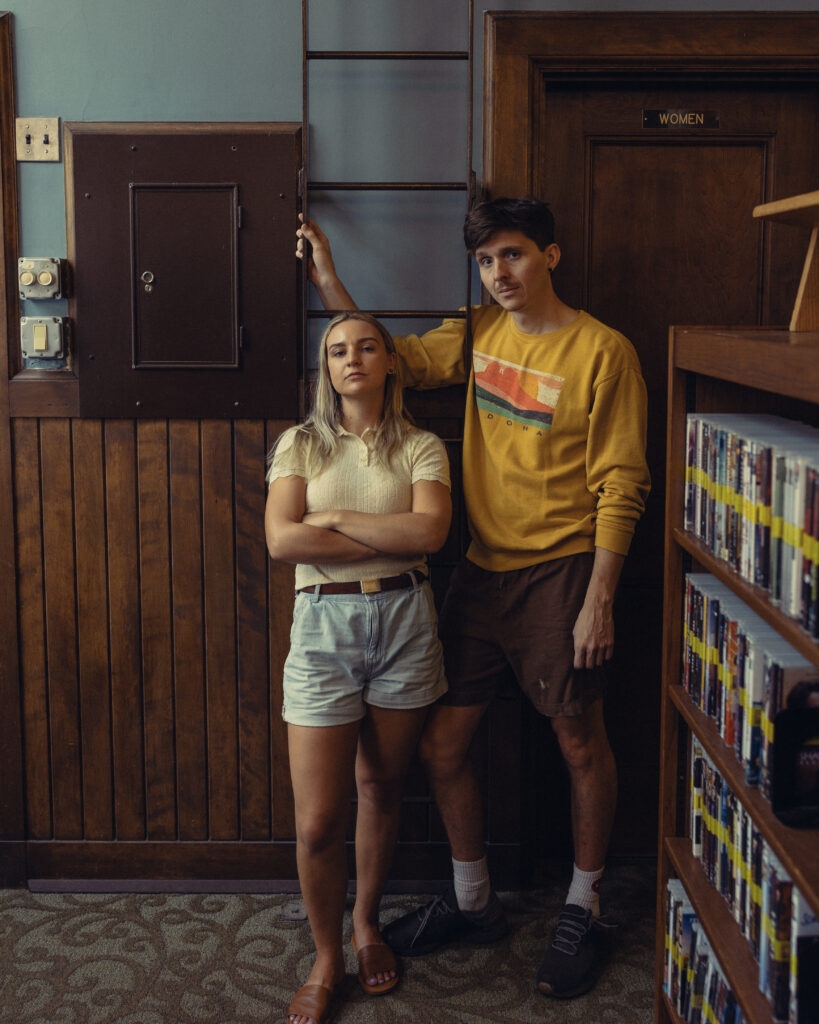 a woman leans against a library ladder as her fiance stands next to her with is arm behind her during a shoot with an engagement photographer