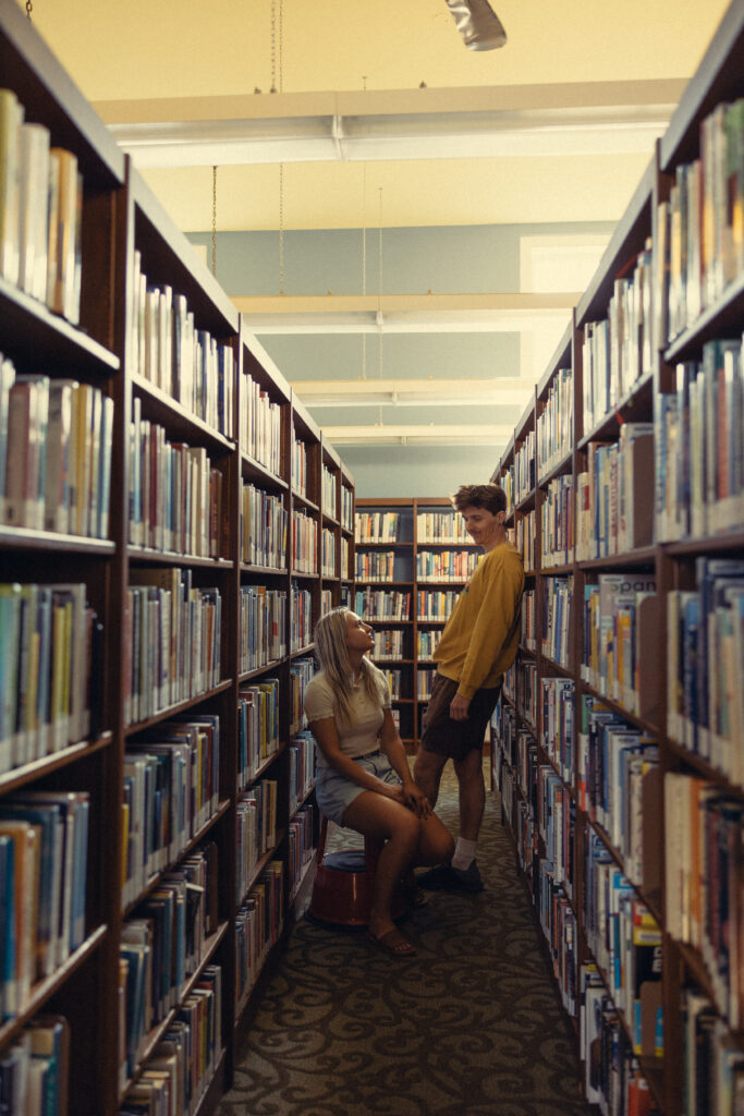 engagement photos grand rapids at a library where a woman sits on a stool and a man leans against a shelf between rows of bookshelves