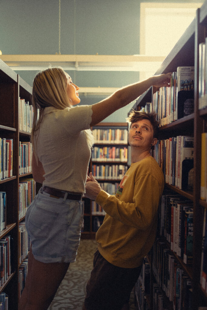 a woman reaches for a book at a library as her fiance leans against the shelf for their engagement photos grand rapids