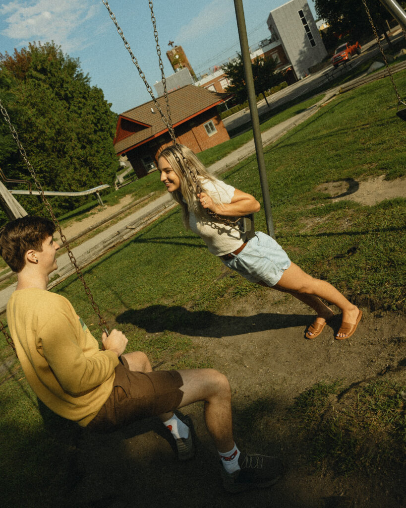 a woman leans while holding on to the swing while looking at her fiance who sits on a swing next to her during engagement photos grand rapids